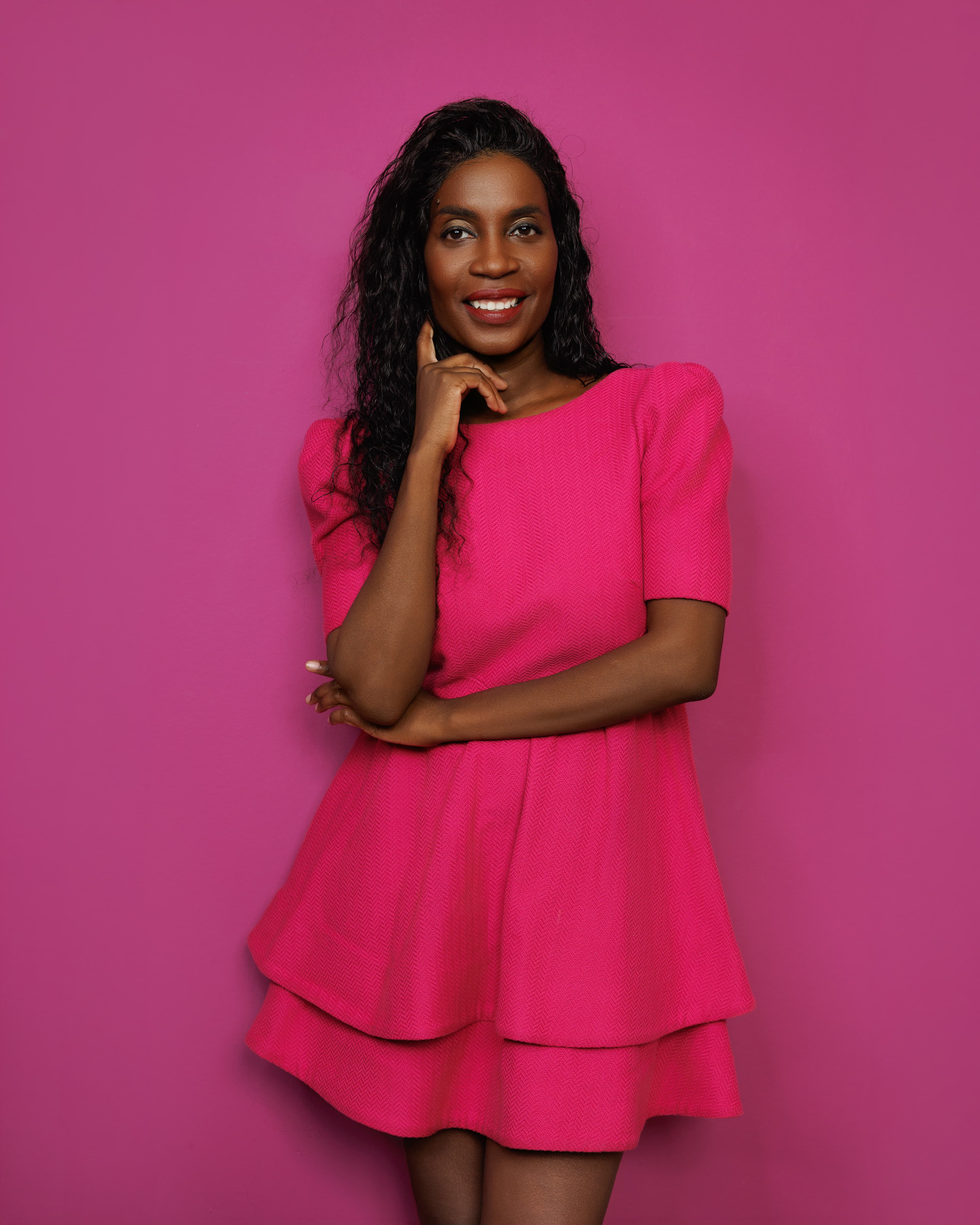 Lächelnde schwarze Frau in einem leuchtend rosa Kleid posiert vor einem farblich passenden Hintergrund.